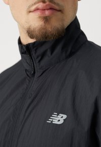 Veste légère noire avec un col haut, arborant un logo New Balance argenté sur la poitrine gauche. Texture lisse et design athlétique.