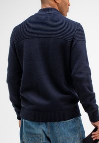 Marineblauwe gebreide trui met een geribde kraag en schouderdetails. Beschikt over een textuur met een zachte afwerking en elastische manchetten en onderkant.