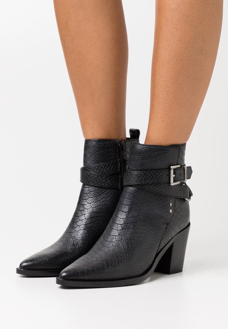 Bottines noires à cheville en similicuir texturé effet peau de serpent, avec bouts pointus, talons blocs et sangles à boucle, portées sur des jambes nues sur fond blanc.
