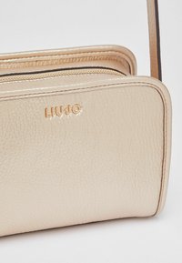 Bolso metálico dorado con superficie texturizada, que incluye una cremallera visible y un logo de la marca en relieve. Diseño compacto y rectangular con una correa para el hombro.