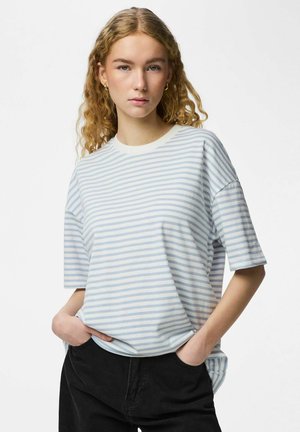 Jeune femme aux cheveux bouclés portant une chemise à manches courtes rayée bleu clair et blanc et un pantalon noir, les mains dans les poches, expression neutre.