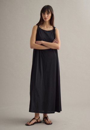 Frau steht mit verschränkten Armen, trägt ein ärmelloses schwarzes Maxikleid und braune Sandalen vor neutralem Hintergrund.