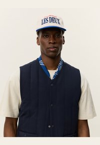 Navy quilted vest with buttons, worn over a light t-shirt. Blue patterned scarf peeks from the collar. White cap with “LES DEUX” text.