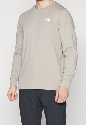 Homme portant un t-shirt à manches longues beige clair avec le logo The North Face sur la poitrine et un pantalon noir.