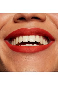 Närbild av en leende mun med klarröd matt läppstift. Tänderna är vita och raka, med synliga detaljer i läpparnas textur och färg.