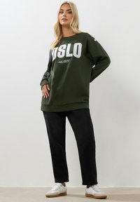 Felpa verde oliva con la scritta "OSLO" e "est. 1978" stampata in bianco. Presenta dettagli a bottoni sulla spalla, abbinata a pantaloni neri e scarpe bianche.