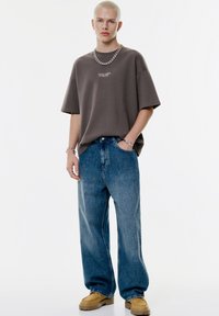 Overdimensioneret brun T-shirt med tekst, parret med løse blå jeans og beige chunky sko. Sølv kæden Halskæde og armbånd tilføjer detaljer.