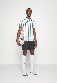 Man in een gestreept voetbaltshirt, zwarte shorts, witte kniekousen en voetbalschoenen die een wit en blauwe voetbal vasthoudt.