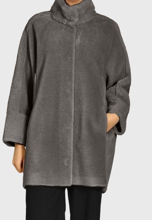 Personne portant un manteau ample en fausse fourrure grise mi-longue avec un col montant et des manches longues, une main dans la poche du manteau, pantalon noir visible.