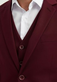 Veste de costume bordeaux avec une chemise blanche en dessous, présentant un col cranté, trois boutons et une poche poitrine sur le côté gauche.