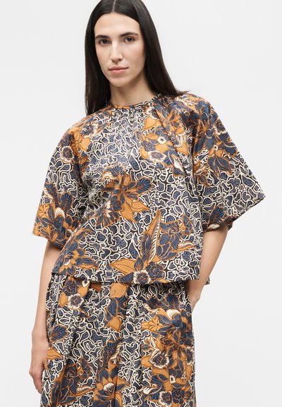 Γυναίκα με μακριά σκούρα μαλλιά που φοράει ένα μαλακό τοπ και παντελόνι με ένα περίπλοκο πορτοκαλί, ναυτικό και λευκό λουλουδάτο σχέδιο.