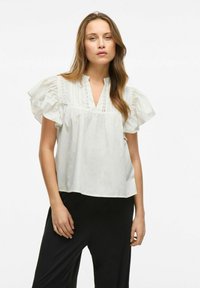 Blusa bianca a maniche corte con maniche arricciate, scollatura a V e dettagli in pizzo. Tessuto morbido con una vestibilità rilassata, abbinata a pantaloni neri.