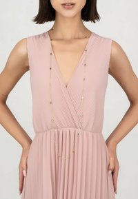 Long gold necklace with colorful beads draped over a pleated blush pink dress featuring a deep V-neck and gathered waist.
