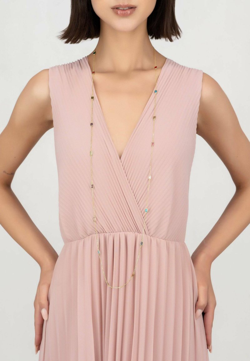 Long gold necklace with colorful beads draped over a pleated blush pink dress featuring a deep V-neck and gathered waist.
