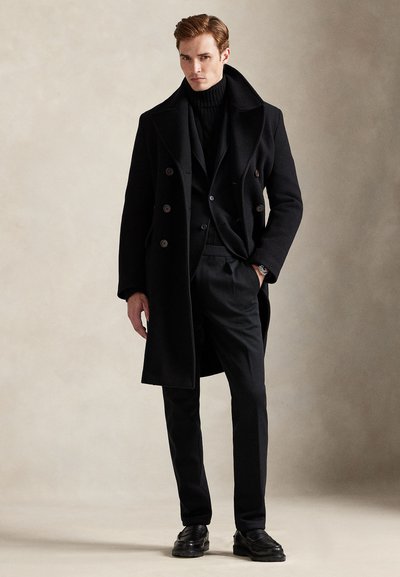 Μαύρο μάλλινο παλτό διπλής σειράς με μεγάλα κουμπιά, φορεμένο πάνω από μαύρο ζιβάγκο και κοστούμι, συνδυασμένο με στενά παντελόνια και μαύρα παπούτσια.