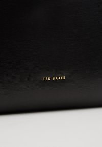 Surface texturée noire avec un petit logo doré "TED BAKER" centré près du bord inférieur.