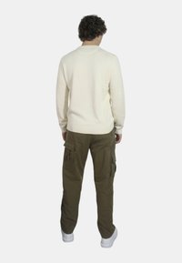 Maglione lavorato a maglia color crema con scollo a girocollo; pantaloni cargo verde oliva con tasche laterali; abbinati a sneakers bianche; visto da dietro.