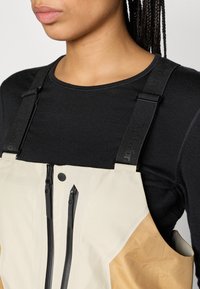 Beige és barna overál fekete cipzárakkal és állítható fekete pántokkal. Sima anyag, oldalsó zsebbel; fekete felső fölött viselve.