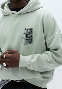 Lichtgroene hoodie gemaakt van zachte stof, met een voorkantgrafiek van gestapelde zwarte blokken met tekst. Geribbelde manchetten en een ontspannen pasvorm.