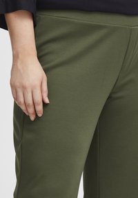 Gröna bomullsblandade byxor med en jämn textur, med en anpassad midja och en sidsöm. Visas med en hand som vilar på tyget.