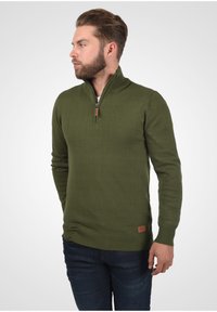 Olivgrüner Zip-Neck-Pullover aus Stricktextur, mit geripptem Kragen und Bündchen, sowie einem Leder-Logopatch am Saum.