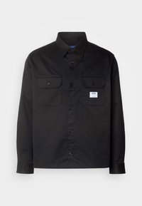 Chemise noire à manches longues en coton avec un devant à boutons, deux poches poitrine et une petite étiquette sur la poche gauche.