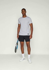 Tricou gri cu mânecă scurtă și pantaloni scurți negri, ținând o rachetă de tenis, stând pe un fundal simplu. Adidași și șosete albe vizibile.
