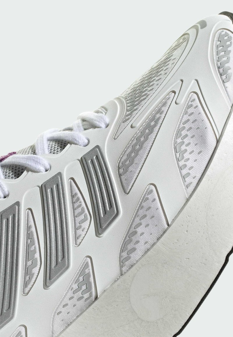 Chaussure de sport blanche avec des superpositions texturées, des panneaux en mesh et une semelle légère. Les accents comprennent des détails gris et des lacets blancs plats.