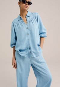 Camisa de denim azul claro con botonera al frente, textura suave y mangas cortas. Combinada con pantalones holgados a juego, que presentan un diseño relajado.