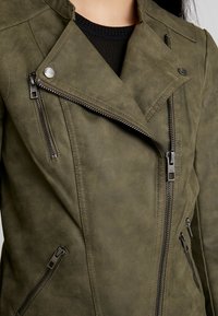 Veste en simili cuir vert olive avec une fermeture éclair asymétrique, des accents métalliques et des poches zippées, dotée d'une finition texturée.