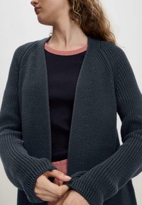 Hiilenharmaa neulecardigan, jossa on avoin etuosa, ribbineulehihat ja teksturoitu pinta. Päällä tummansininen toppi, jossa on kontrastina vaaleanpunainen pääntie.