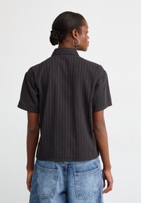Svart kortermet skjorte med hvite striper, skreddersydd design, krage, noe kortere i lengden, vist fra baksiden med blå denimshorts.