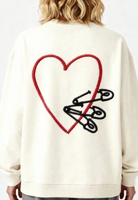 Felpa color crema con un grande cuore ricamato rosso e spilli di sicurezza neri in un design a strati sul retro. Tessuto morbido e vestibilità rilassata.