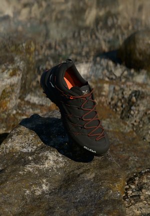 Čierne a oranžové turistické topánky s šnúrkami ležiace na skalnatom teréne, s logom "SALEWA" na oblasti prstovej časti.