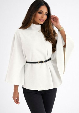 Femme aux longs cheveux bruns portant un haut-cape blanc à col montant avec de larges manches, ceinture noire, pantalon noir, et une montre en or au poignet gauche.