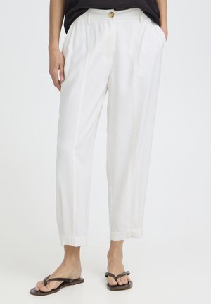 Witte op maat gemaakte broek met omgeslagen pijpen, zijzakken en een bruine knoop in de taille, gecombineerd met zwarte teenslippers.