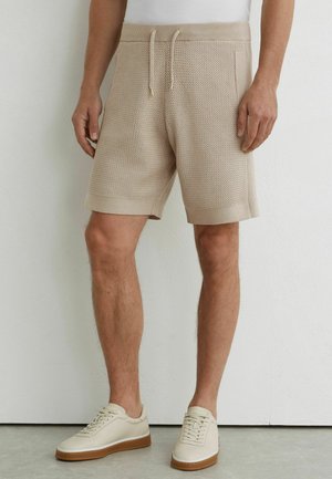 Mann trägt beige gestrickte Shorts mit Kordelzug und cremefarbenen Turnschuhen, steht vor einer weißen Wand und einem grauen Boden.