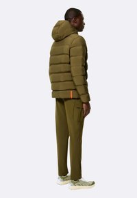 Giacca a vento verde oliva con motivo a righe orizzontali e cappuccio, abbinata a pantaloni cargo coordinati. I dettagli includono rifiniture arancioni e sneakers testurizzati.
