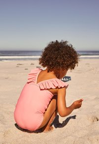 Rožinė vientisa maudymosi apranga su raukiniais ant peties. Vaikas gūžteli smėlyje, žaidžia su šviesiai mėlynu kibirėliu, fone matyti paplūdimio scena.