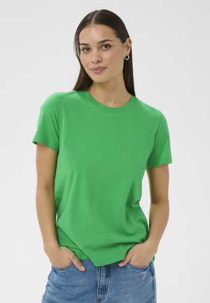 Kvinde med langt mørkt hår iført guld øreringe, en skriggrøn kortærmet t-shirt og blå denimjeans med hænderne i lommerne.