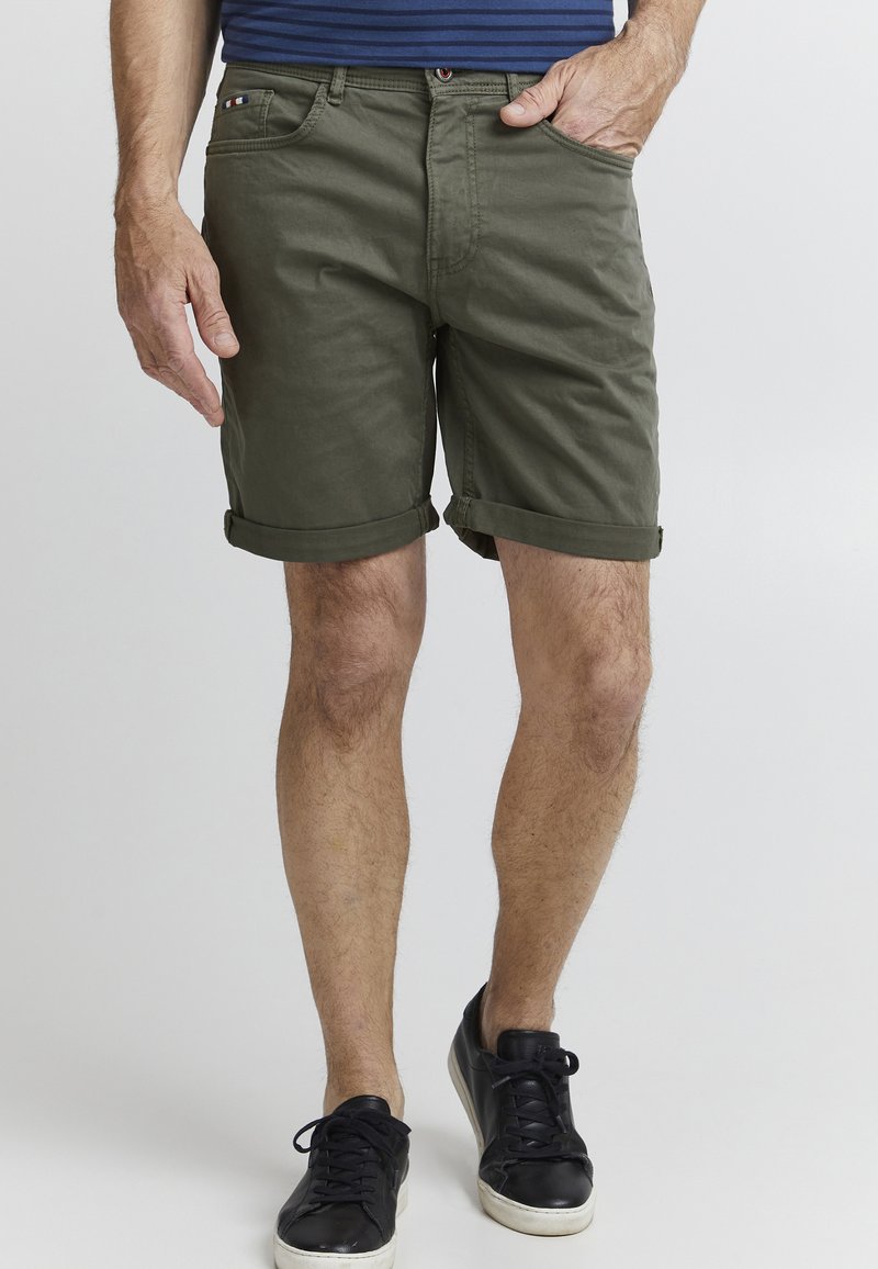 Olivenfargede bomullsshorts med knapp- og glidelåslukking, oppbrettede ermer, sidelommer og et merketikett på linningen. Svarte joggesko er brukt.