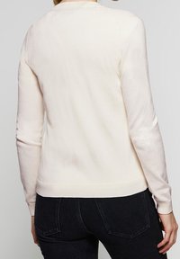 Krémově zbarvený lehký svetr s dlouhými rukávy, kulatým výstřihem a hladkou, měkkou texturou. Má přiléhavý střih a jemné švové detaily.