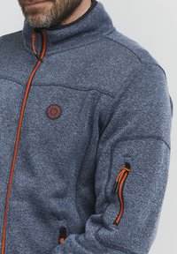 Tumedast sinisest fleece-jakist tekstuursed materjalid, oranž tõmblukk ja aktsendid, kõrge krae ning vasakul varrukal tõmbluku tasku.