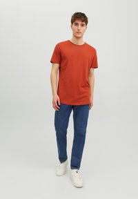 T-shirt a maniche corte color ruggine, colletto rotondo, abbinato a jeans in denim blu e sneakers bianche. Design semplice e tessuto leggero.