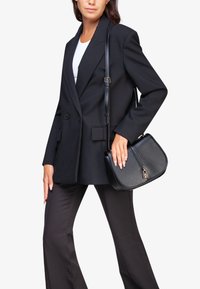 Blazer nero doppiopetto con revers abbinato a pantaloni neri a zampa. La modella tiene una borsa nera strutturata con hardware argentato.