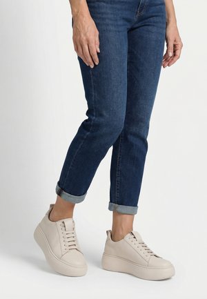 Persoon die opgerolde blauwe spijkerbroek en lichtbeige platformsneakers draagt, met de voeten iets uit elkaar staand op een witte achtergrond.