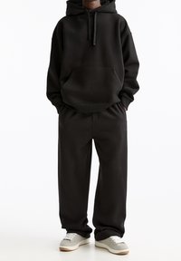 Zwarte hoodie van zachte stof met een voorzak en een capuchon met trekkoord, gecombineerd met bijpassende sweatpants in een losse pasvorm. Grijze sneakers maken de look compleet.