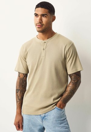 T-shirt henley beige à côtes à manches courtes avec patte de boutonnage à trois boutons ; porté avec un denim bleu clair, montrant des tatouages sur le bras.