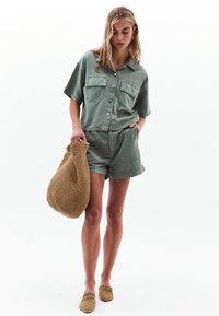 Lichtgroen shirt met korte mouwen en twee voorzakken, gecombineerd met bijpassende shorts. Een geweven bruine tas en open teen schoenen maken de outfit compleet.