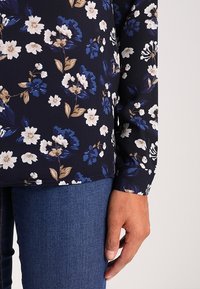 Blusa floreale con sfondo blu navy, caratterizzata da fiori bianchi, blu e beige. Maniche lunghe e un design aderente, indossata sopra jeans di denim.